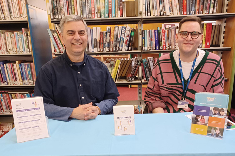 Two Reconnect staff members smiling to camera sitting behind a People Know How stall at a community library