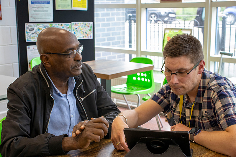 A People Know How VIP supporting someone to use a device as part of a Reconnect digital group, the two sit side by side and look at a tablet