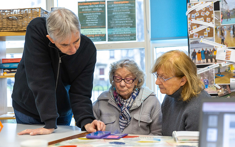 Three people at a Reconnect digital group looking at a device, two are sitting and one is standing